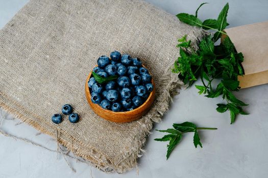 A small wooden bowl with berries. Juicy and ripe blueberries in an eco-friendly dish. Sprigs of fresh mint in a kraft bag. The concept of healthy eating and nutrition. Drops of water on blueberries.