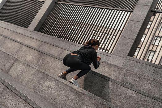 Young woman doing leg exercises at stone stairs outside with city on background. Beautiful cauceuropean woman jumping over big step, squat exercise. Fitness and health concepts. Rear view