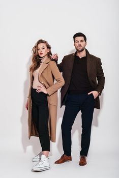 Full-length portrait of couple. Woman in beige coat, blue trousers and white converse and man wearing blue classical trousers with brown jacket. Isolated over grey background. Couple goals. Happy together