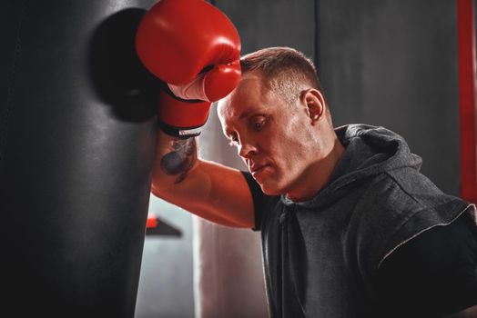 Tired young man in sports clothing training hard on heavy punch bag. Muscular sportsman with red boxing gloves looking away while standing in boxing gym