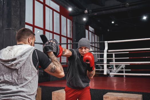 Training hard. Confident muscal athlete in red gloves training on boxing paws with partner in black boxing gym