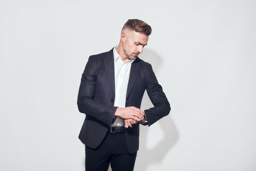 Studio shot of handsome bearded businessman in classic suit looking at watch on his hand while standing against grey background. Time concept. Business look.