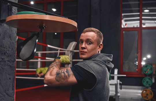 Hard arm workout. Muscular young sportsman with green hands wraps punching speed bag while training his boxing skills in colored gym