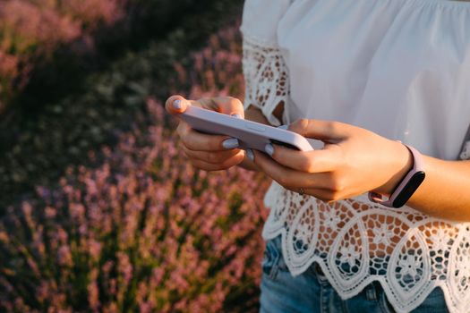 A girl in a white cotton top with lace holds a smartphone in a purple case. Beautiful lavender field. Modern technologies. Fitness bracelet with purple silicone strap. Waterproof smart watch.