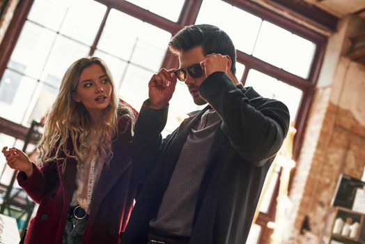 Wrong size of sunglasses. Happy young joyful couple choosing sunglasses for man at small street market. Young man wearing sunglasses and ask opinion of his girlfriend