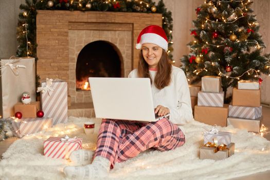 Beautiful young female in santa hat celebrating christmas and new year at home, using laptop, posing on background of christmas tree, fireplace, present boxes and lights.