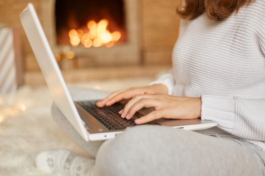 Faceless woman typing message on her laptop computer as she relaxes near burning warm fire, unknown female working online while sitting on floor with crossed legs, holding hotebook on her knees.