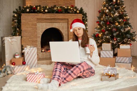 Christmas at home, celebrating winter holidays during quarantine, lady with sad facial expression sits on floor with notebook on knees, drinking coffee or tea, holding cup in hands.