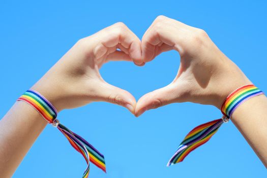 Close up of Woman hands in a rainbow bracelet making a heart shape form on blue sky background. High quality photo.