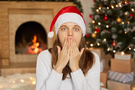 Young European woman wears santa hat and white sweater, being at home around christmas decoration, covers mouth with hands, being very surprised, pose with fireplace and x-mas tree on background