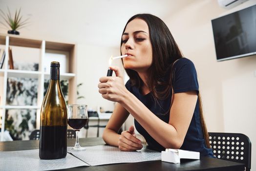 Portrait of young woman in despair, smoking cigarette while sitting at table with glass of alcohol at home. Female alcoholism concept. Protest in the treatment of alcohol addiction.