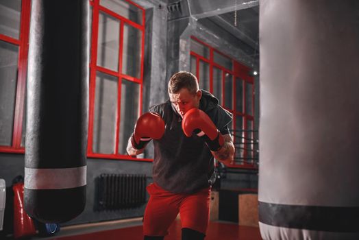 Handsome muscular sportsman in sports clothing boxing on heavy punch bag. Hard training on boxing gym background