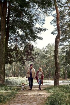 Young family couple are walking in woods with their dog. Stylish man is looking at his girlfriend while holding her hand. Bright sun is shining throw the trees, outumn season. Full length