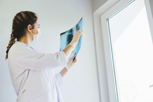 A radiologist in a medical mask and gloves radiates an x-ray image of the patient's lungs. The girl doctor works on the front line. The doctor is holding an X-ray of his lungs.