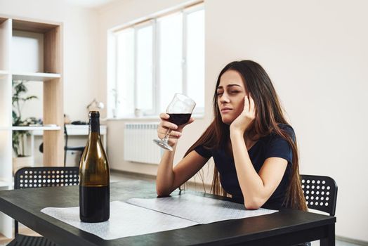 Depressed, divorced woman sitting alone in kitchen at home and holding a glass of red wine because of problems at work and troubles in relationships. Social and life problems
