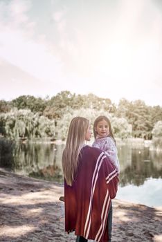 A little curly girl in sweater is hugging her mother in woods near lake. She is wearing pink sweater and young woman is in red knitted coat.