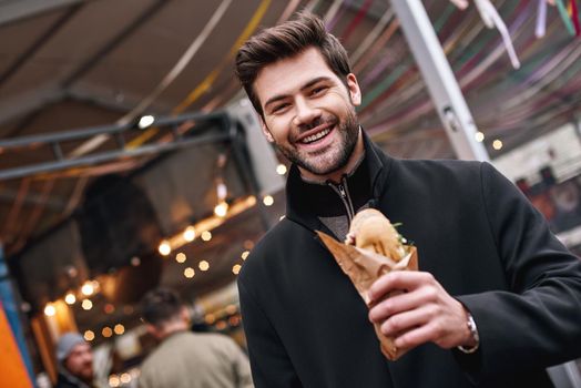 Happy to eat sandwich. Young stylish man in black coat is eating out sandwich at sandwich bar at street fair.