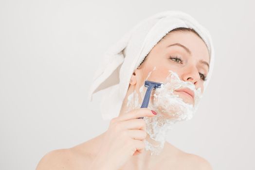 Cheerful caucasian woman with a towel on her head and shaving foam on her face holds a razor on a white background.