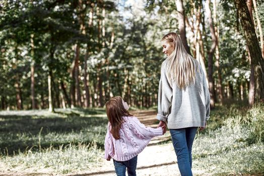 A little curly girl in sweater is walking with her mother in woods. Cold season, bright sun is seen through the trees.