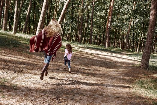 A little curly girl in sweater is running with her mother in woods. Cold season, bright sun is seen through the trees. She is wearing pink sweater and young woman is red knitted coat. Rear view