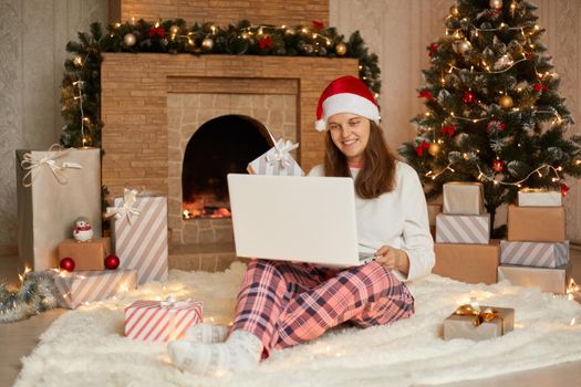 Photo of cute charming attractive girl working in eve of new year with her laptop or having video call for greeting somebody, wearing checkered pants and santa hat, looks at notebook scree with smile.