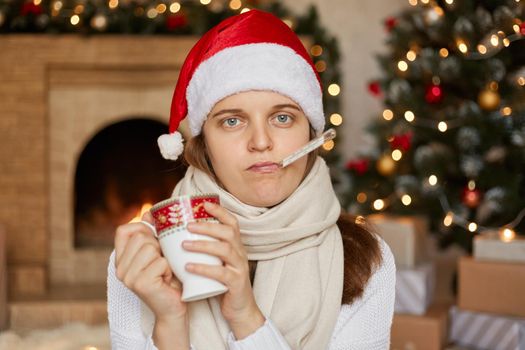 Young ill woman in santa hat, wrapped in scarf, measuring temperature and holding cup of warming drink, looks at camera with unhappy facial expression, posing indoor with fireplace and x-mas tree .