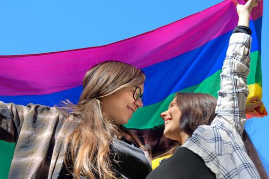 Affectionate lesbian couple holding an LGBT flag under blue sky. High quality photo.