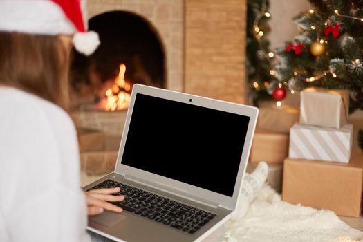 Photo of charming lady sitting on floor and holding laptop with blank screen, wear red santa claus hat and white sweater, posing in decorated x-mas living room. Copy space.