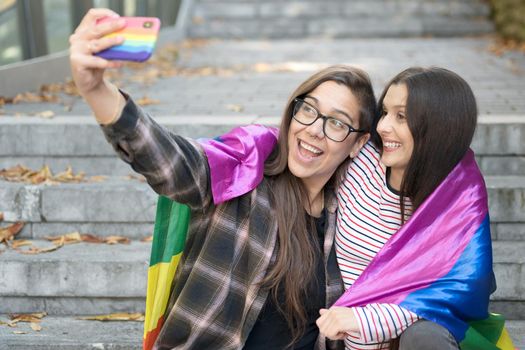 Cheerful happy lesbian couple holding lgbt flag taking a selfie outdoors. High quality photo