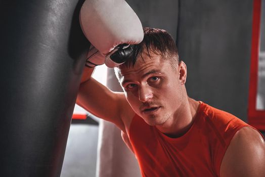 Tired young man in sports clothing training hard on heavy punch bag. Muscular sportsman with white boxing gloves looking away while standing in boxing gym