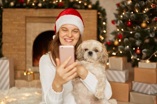 Pretty cheerful woman poses with her Pekingese dog and taking selfie, spend winter holidays at home, smiling woman wit smart phone in hands sitting on floor in festive living room.