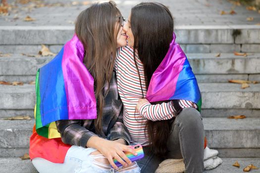 Lesbian couple sitting on steps with rainbow flag, hugging and kissing at urban scenery. High quality photo.