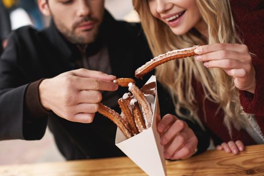 Couple are eating outdoors . Young couple are eating churros at the street food market. Cold season. Close-up photo of couple eating from one package