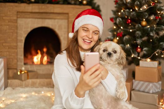 Woman and dog in sweater having fun takes selfie portrait on smart phone or have video call with somebody, enjoying Christmas time at home, wearing santa claus hat, posing in festive living room.