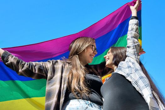 Affectionate lesbian couple holding an LGBT flag under blue sky. High quality photo.