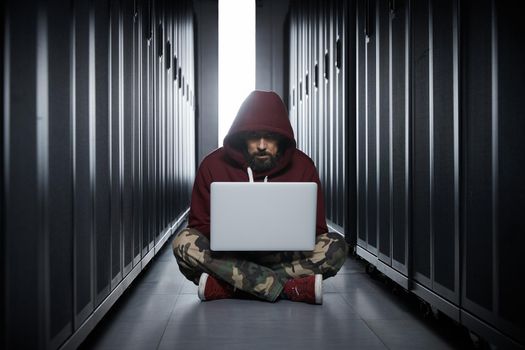 The hacker in a hood sits with the computer among servers, the burglar, face covered with a hood. Storage of data, cloudy services, system breaking, attack of hackers. High quality photo