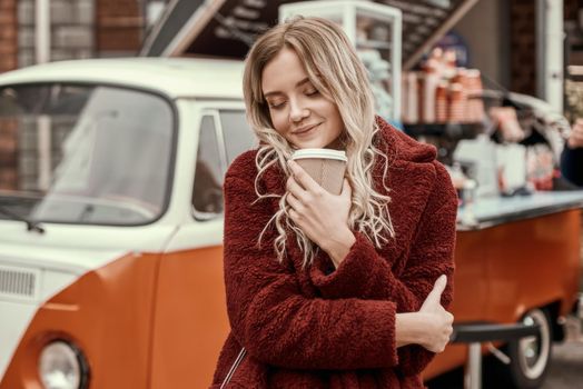 So aromatic coffee. Blonde beautiful white woman with long hair sniffs coffee and huggs herself on urban background. Outdoor photo of lovable girl in red eco fur coat standing with cup of latte near car.