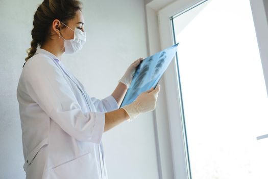 A radiologist in a medical mask and gloves radiates an x-ray image of the patient's lungs. The girl doctor works on the front line. The doctor is holding an X-ray of his lungs.