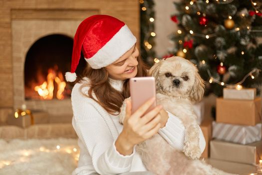 Photo of smiling lady with dog at Christmas, posing in festive room near fireplace and taking selfie or having video call, lady looking at her pet with smile and holding phone in hands.
