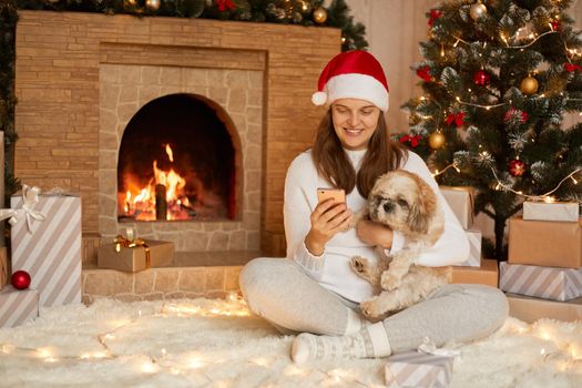 Smiling lady sitting on floor with smart phone and hugging her pekingese dog, lady checking social network via mobile phone, sits with crossed legs, looks smiling at device's screen.