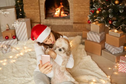 Beautiful young woman in Christmas hat and sweater with small dog hugs, sitting in festive living room on floor near fireplace, lady keeps lips rounded wants to kiss pekingese puppy.