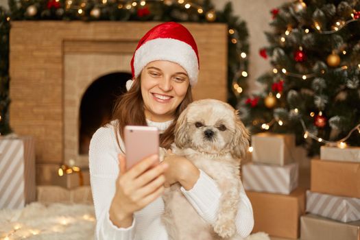 Girl making selfie with nice puppy while sitting in festive room with fireplace and x-mas tree, girl looks at mobile phones's screen with charming smile, wearing santa hat and casual jumper.