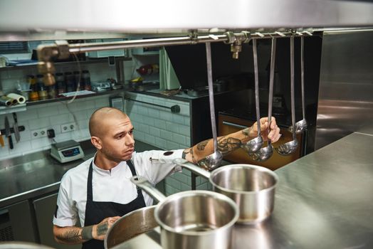 Cooking soup. Young male chef in apron choosing a ladle for his famous soup while standing in a restaurant kitchen. Cooking concept