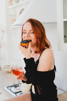 The girl is sitting eating a cake and drinking wine. A young girl in a black negligee is sitting in the kitchen eating a cake and drinking wine. Bright kitchen. Sexy girl is resting.