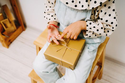 A little girl in a denim jumpsuit sits on a wooden chair and holds a gift box on her knees.