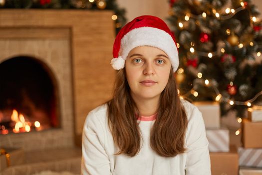 Christmas woman in santa hat sitting in decorating living room near fireplace and fir tree, looks at camera, wearing casual shirt, has calm facial expression.