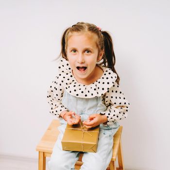 Funny girl sits on a wooden stool with a gift on her knees. A little girl in a polka dot print shirt. The child smiles.