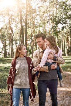 Young parents with little daughter in autumn forest. Father is holding baby girl, mother walks nearby and touching her hand at the left side