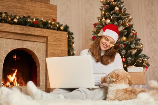 Young happy freelancer working at home while sitting on floor with her pekingese dog, posing in festive room decorated with x-mas lights, sits near fireplace, looks smiling at her pet.