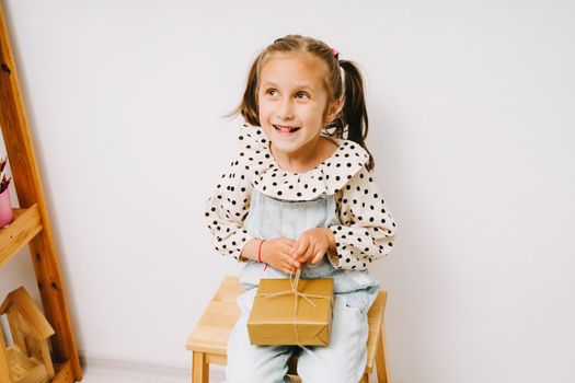 A little girl untied a bow on her gift. A little girl in a denim jumpsuit sits on a wooden chair and holds a gift box on her knees. The girl smiles slyly.